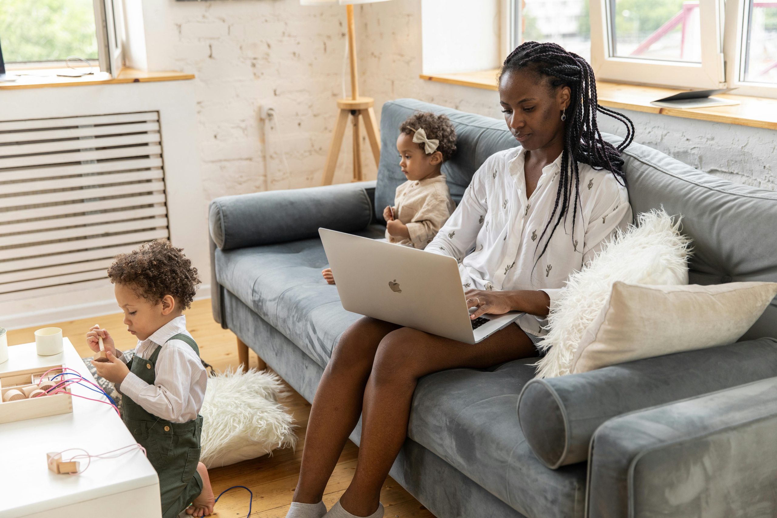 A mother works from home on her laptop while two children play nearby on a couch.
